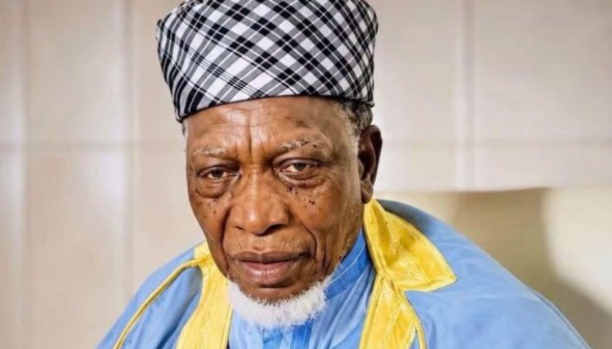 Portrait of an elderly man with a black-and-white plaid head wrap, wearing blue and yellow traditional clothing, looking at the camera.