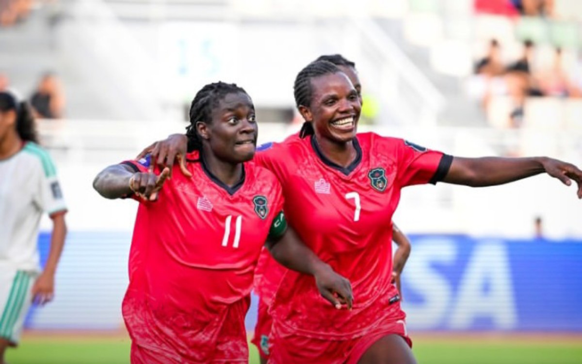 Two female soccer players in red uniforms celebrate together on the field, arms around each other and smiling (numbers 11 and 7 visible).