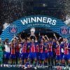 Football team in blue-red kits celebrate on the field after winning, lifting the trophy under a 'WINNERS' banner with confetti filling the air.