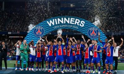 Football team in blue-red kits celebrate on the field after winning, lifting the trophy under a 'WINNERS' banner with confetti filling the air.