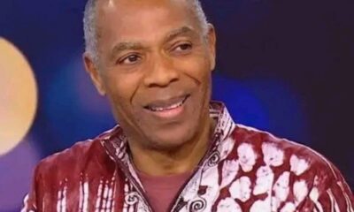 Smiling man with gray hair wearing a red and white patterned shirt on a stage background.