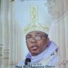 Catholic bishop speaking into a microphone, wearing an ornate white and gold mitre and purple vestments, with church columns in the background (text reads 'His Grace, Most Rev. Augustine Obiora Akubeze')