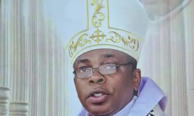 Catholic bishop speaking into a microphone, wearing an ornate white and gold mitre and purple vestments, with church columns in the background (text reads 'His Grace, Most Rev. Augustine Obiora Akubeze')