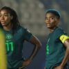 Two female soccer players in green jerseys stand with hands on hips during a match, one wearing number 11 and the other number 3 with a yellow captain's armband, stadium lights in the background.