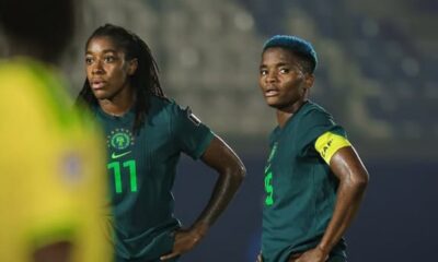 Two female soccer players in green jerseys stand with hands on hips during a match, one wearing number 11 and the other number 3 with a yellow captain's armband, stadium lights in the background.