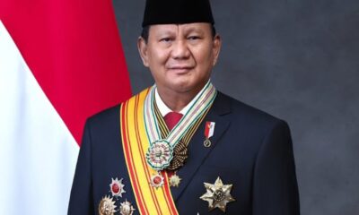 Formal portrait of a man in a dark suit with multiple medals and a striped sash, standing in front of a red and white flag.