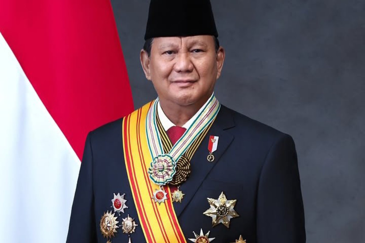 Formal portrait of a man in a dark suit with multiple medals and a striped sash, standing in front of a red and white flag.