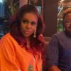 Two adults sit on a couch in a dim restaurant; woman wears an orange top with red hair and long earrings, man beside her wearing glasses.
