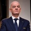 Bald man in a navy suit and blue tie with a FIFA emblem on the lapel, looking serious at a formal event