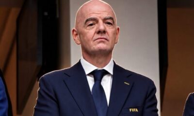 Bald man in a navy suit and blue tie with a FIFA emblem on the lapel, looking serious at a formal event