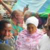 Crowd of people at a public event, a woman in a white headscarf and pink floral outfit in the center among others nearby.