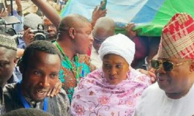 Crowd of people at a public event, a woman in a white headscarf and pink floral outfit in the center among others nearby.