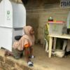 Woman in a beige hijab kneels on a step beside a tall white voting booth at a polling station, as staff stand at a table nearby.