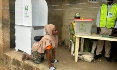 Woman in a beige hijab kneels on a step beside a tall white voting booth at a polling station, as staff stand at a table nearby.