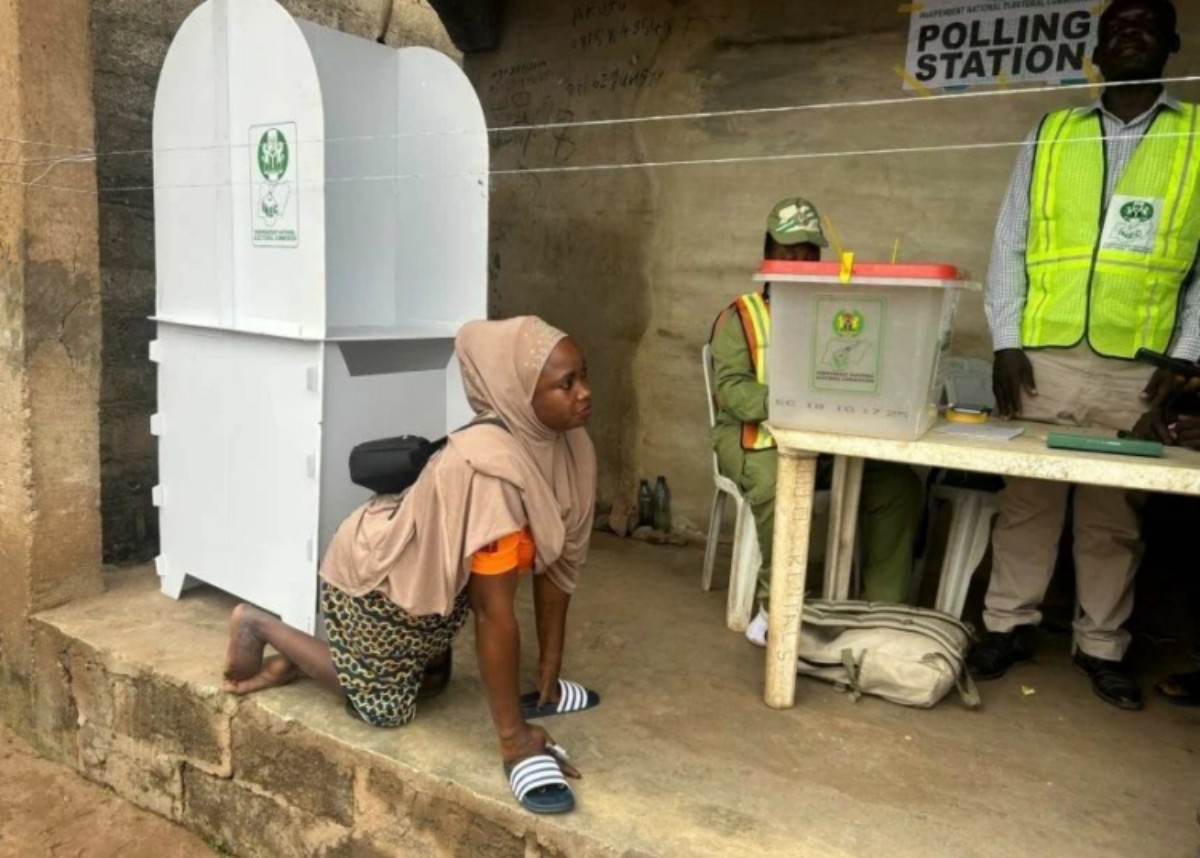 Woman in a beige hijab kneels on a step beside a tall white voting booth at a polling station, as staff stand at a table nearby.