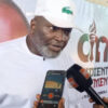 Man in a white traditional outfit and cap being interviewed, with a reporter holding a smartphone showing a voice recorder app and a black microphone nearby; backdrop features a logo and text.