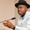 Man wearing a black hat speaks into a microphone at a meeting, gesturing with one hand while seated at a desk.