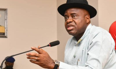 Man wearing a black hat speaks into a microphone at a meeting, gesturing with one hand while seated at a desk.
