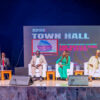 Panel discussion on a stage with six people seated in white chairs, a large screen behind reads 'TOWN HALL' and 'Arise News'.