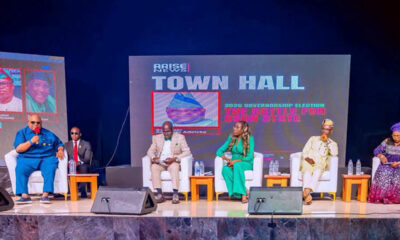 Panel discussion on a stage with six people seated in white chairs, a large screen behind reads 'TOWN HALL' and 'Arise News'.