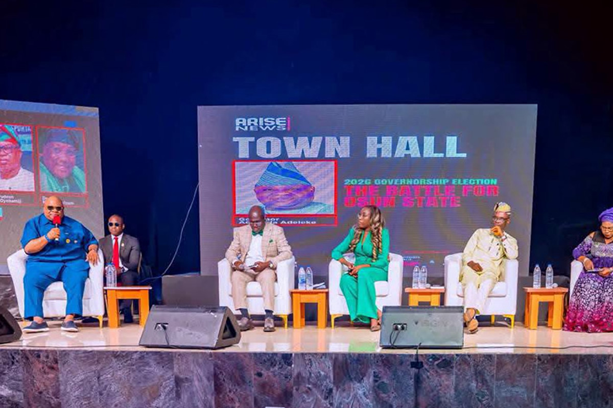 Panel discussion on a stage with six people seated in white chairs, a large screen behind reads 'TOWN HALL' and 'Arise News'.
