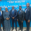 Five professionally dressed individuals posing for a group photo in front of a bright blue wall with a quote mural. (woman in blue patterned dress, others in suits)