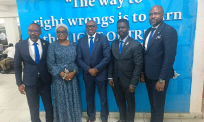 Five professionally dressed individuals posing for a group photo in front of a bright blue wall with a quote mural. (woman in blue patterned dress, others in suits)