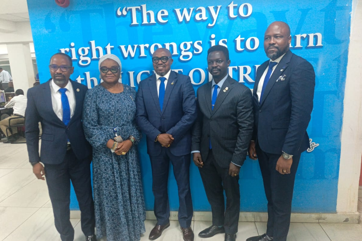 Five professionally dressed individuals posing for a group photo in front of a bright blue wall with a quote mural. (woman in blue patterned dress, others in suits)