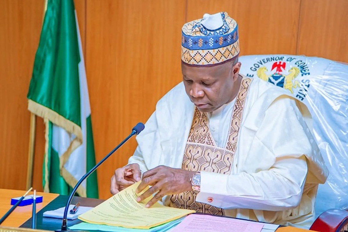 A Nigerian official in traditional attire signs a document at a desk with a microphone nearby.