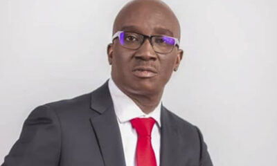 Professional head-and-shoulders portrait of a man in a dark suit and red tie wearing glasses against a light gray background.