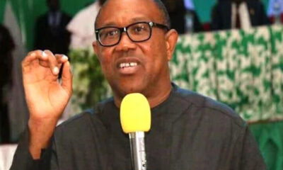 Man wearing glasses speaks into a yellow microphone, gesturing with his right hand during a formal event.