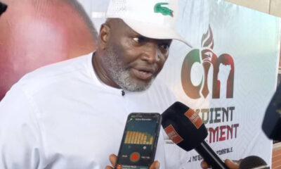 Man in a white traditional outfit and cap being interviewed, with a reporter holding a smartphone showing a voice recorder app and a black microphone nearby; backdrop features a logo and text.