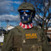 Police special response team member in tactical gear, star-pattern scarf, helmet, and sunglasses outdoors.