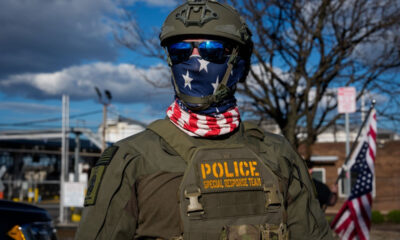 Police special response team member in tactical gear, star-pattern scarf, helmet, and sunglasses outdoors.