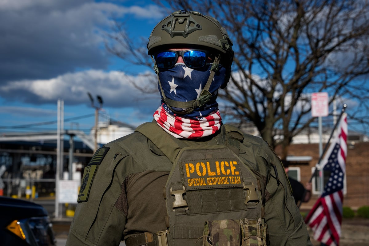 Police special response team member in tactical gear, star-pattern scarf, helmet, and sunglasses outdoors.