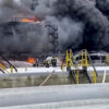 Industrial facility with a large fire and thick black smoke, firefighters in gear standing near yellow safety rails and equipment in the foreground.