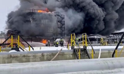 Industrial facility with a large fire and thick black smoke, firefighters in gear standing near yellow safety rails and equipment in the foreground.