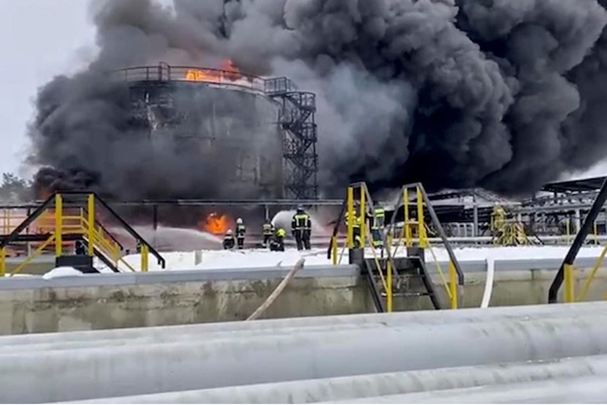 Industrial facility with a large fire and thick black smoke, firefighters in gear standing near yellow safety rails and equipment in the foreground.