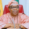 Older Nigerian official in pink traditional attire and cap, seated at a desk with a microphone and red chair behind him.