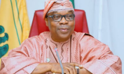 Older Nigerian official in pink traditional attire and cap, seated at a desk with a microphone and red chair behind him.