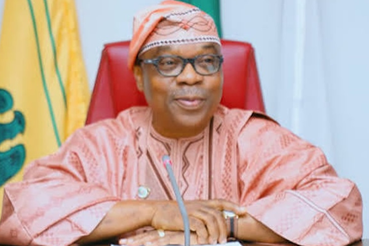 Older Nigerian official in pink traditional attire and cap, seated at a desk with a microphone and red chair behind him.