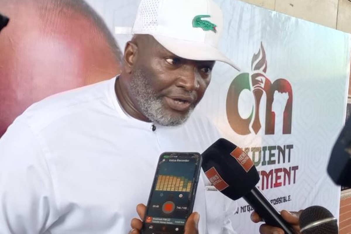 Man in a white traditional outfit and cap being interviewed, with a reporter holding a smartphone showing a voice recorder app and a black microphone nearby; backdrop features a logo and text.