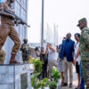 Soldiers guard a memorial as civilians observe a ceremony, with one person taking photos and others watching respectfully.