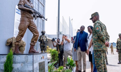 Soldiers guard a memorial as civilians observe a ceremony, with one person taking photos and others watching respectfully.