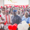 Group of people at a ribbon-cutting ceremony with red and white balloons in the background, some holding a wrapped gift.