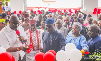 Group of people at a ribbon-cutting ceremony with red and white balloons in the background, some holding a wrapped gift.