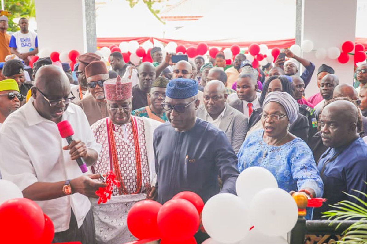 Group of people at a ribbon-cutting ceremony with red and white balloons in the background, some holding a wrapped gift.