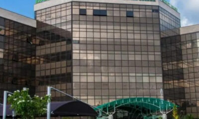 Glass-front office building with reflective panels and a green arched walkway to the entrance.