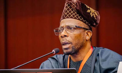 Man wearing a patterned traditional cap and glasses speaks into a microphone at a podium during a formal event.