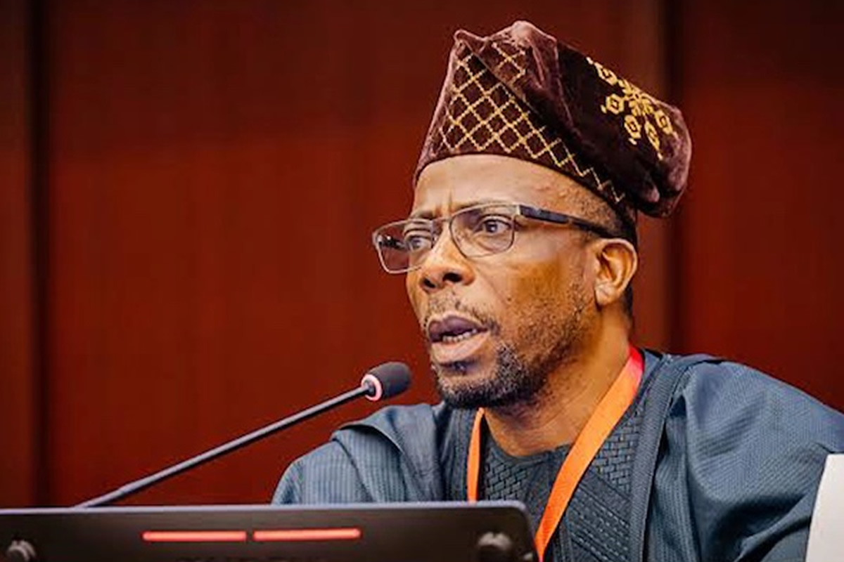 Man wearing a patterned traditional cap and glasses speaks into a microphone at a podium during a formal event.